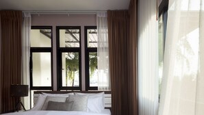 A hotel room with a large bed, a nightstand with a lamp, and a view through a window with curtains. - Nan Garden House (Nan)