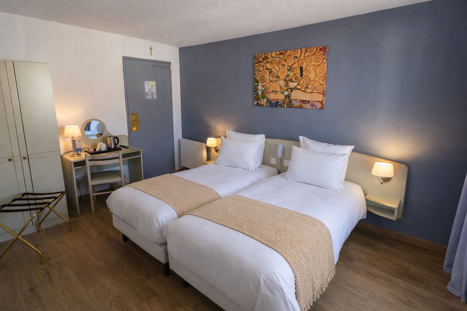 Ein Doppelbett mit einer beige farbenen Decke, ein Schreibtisch mit einer Lampe, ein Spiegel und eine Wand mit einem Gemälde.