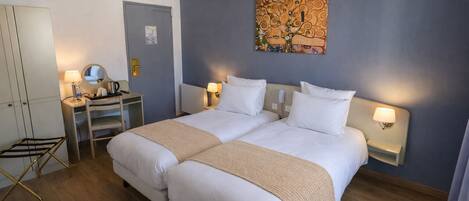 A double bed with a beige blanket, a desk with a lamp, a mirror, and a wall with a painting.
