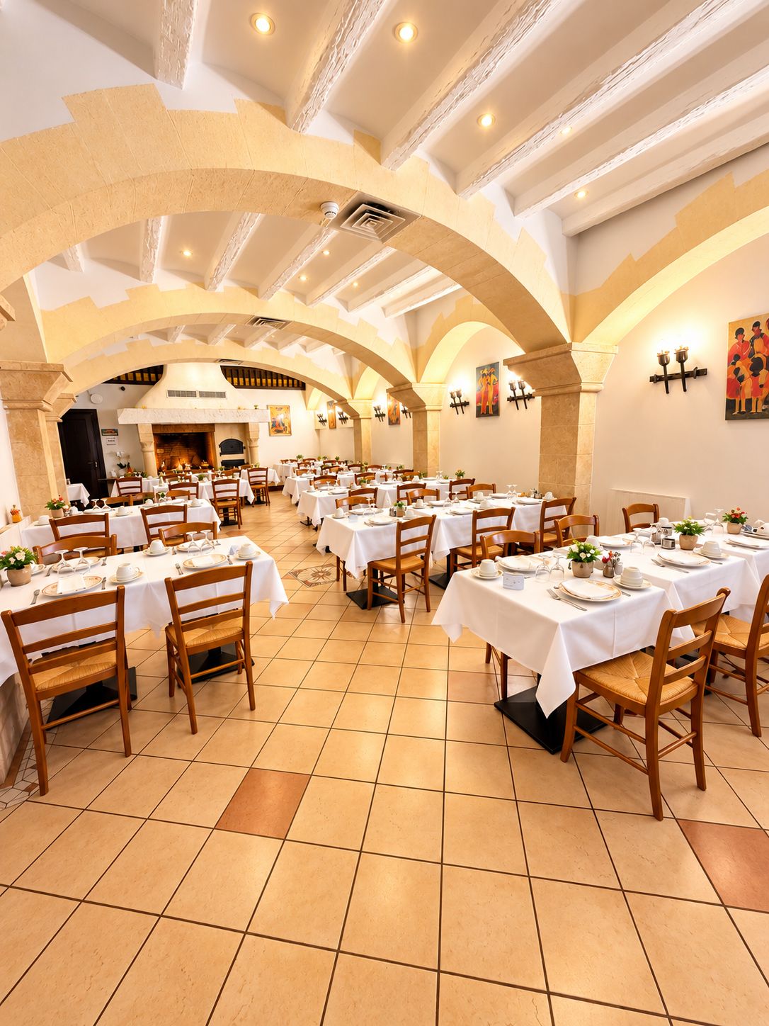 A spacious dining area with multiple tables set for guests, featuring a high ceiling with exposed beams and a central fireplace.