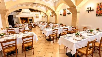 A spacious dining area with multiple tables set for guests, featuring a high ceiling with exposed beams and a central fireplace.