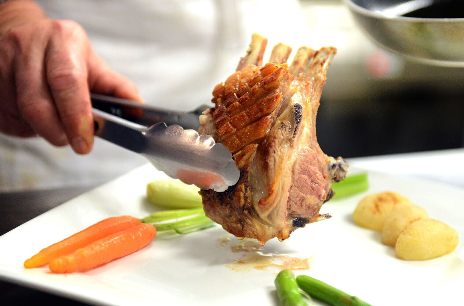 A chef using tongs to handle a cooked rack of lamb on a white plate, accompanied by vegetables.