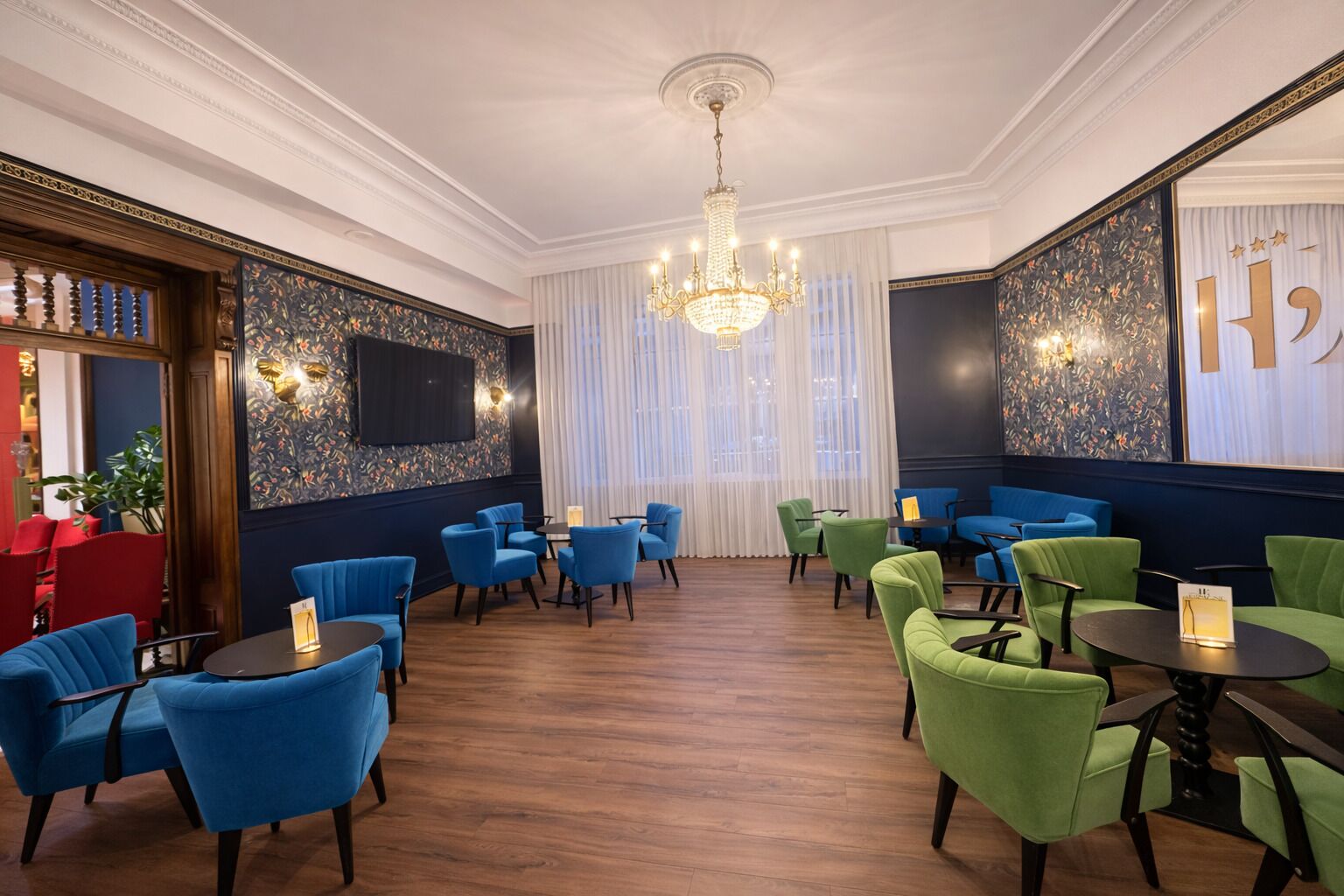 A room with blue and green upholstered chairs, a chandelier, and a large window with sheer curtains.
