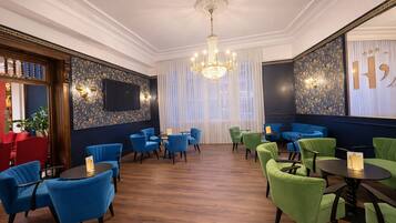 A room with blue and green upholstered chairs, a chandelier, and a large window with sheer curtains.