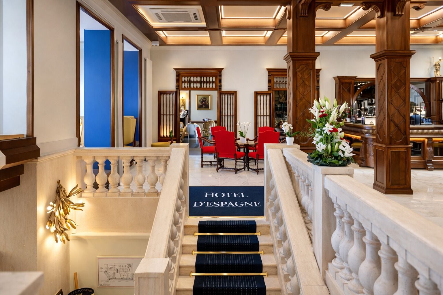 A hotel lobby with a staircase leading to a reception area, wooden columns, and a blue wall.