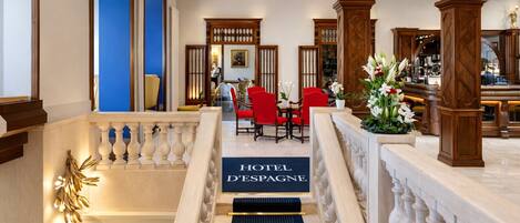 A hotel lobby with a staircase leading to a reception area, wooden columns, and a blue wall.