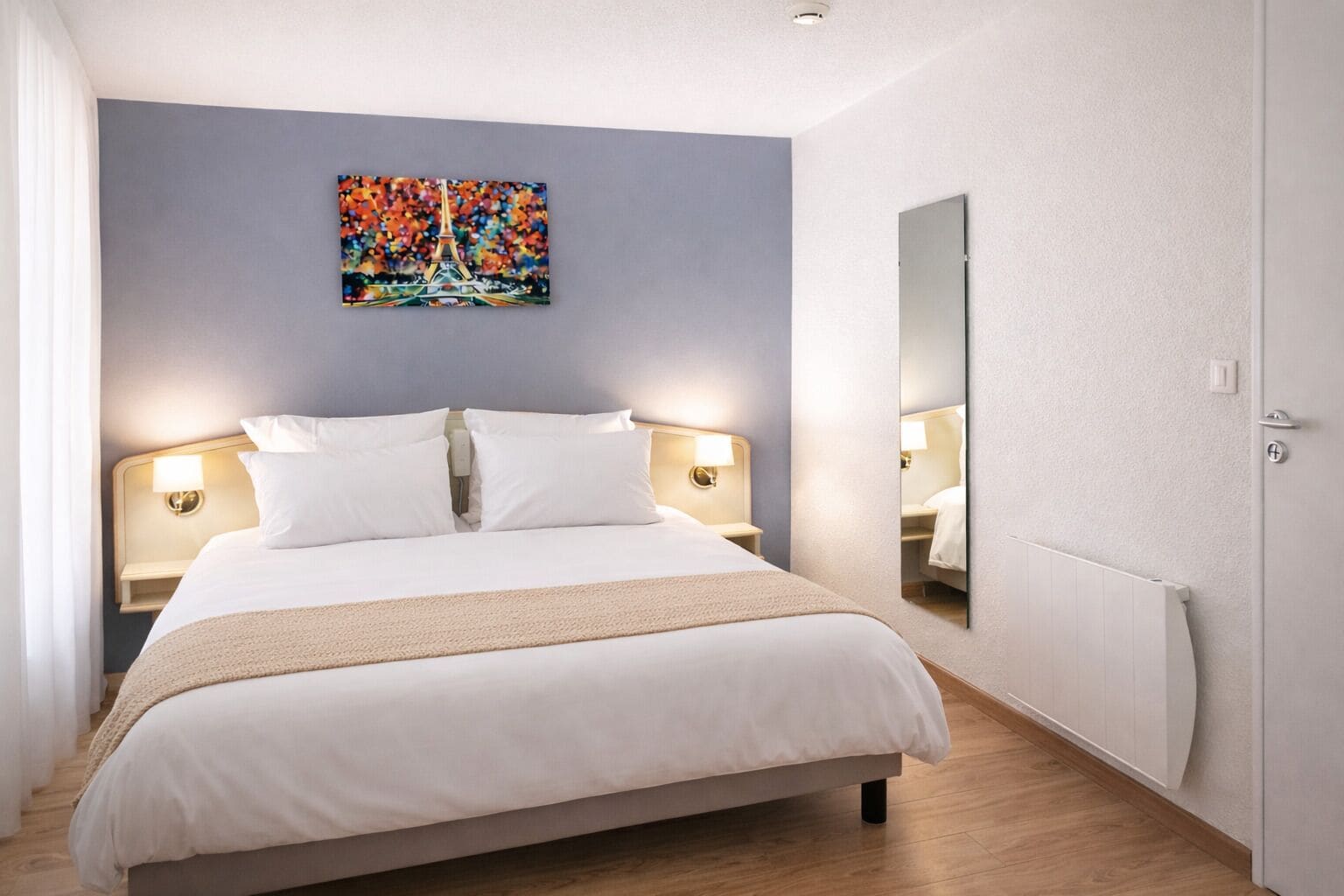 A hotel room with a bed, two bedside lamps, a large mirror, and a painting of the Eiffel Tower.