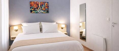 A hotel room with a bed, two bedside lamps, a large mirror, and a painting of the Eiffel Tower.
