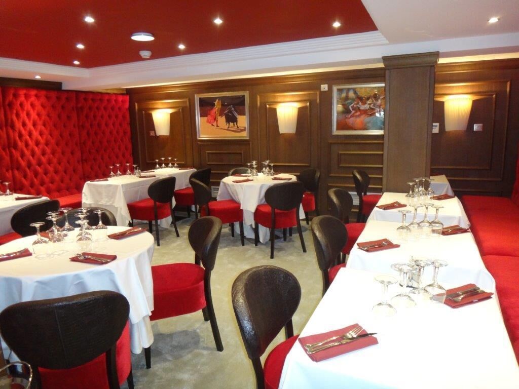 A dining area with multiple round tables, each set with white tablecloths, red chairs, and neatly arranged cutlery and glassware.