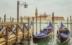 A wooden pier with gondolas docked, a lamp post, and a historic building with domes in the background.