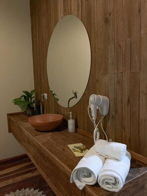 Une coiffeuse en bois avec un miroir rond, un lavabo en forme de bol, un sèche-cheveux et des serviettes.