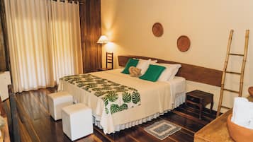 Un dormitorio con piso de madera, cama con cubrecama de hojas verdes, almohadas blancas, lámpara de noche y una escalera de madera.