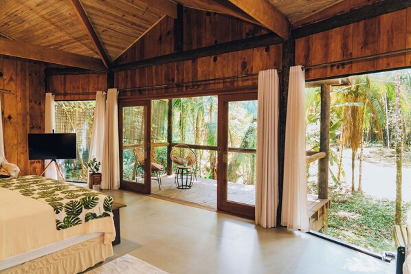 A spacious room with a large bed, a television, and a view of a tropical garden through large glass doors. - Glamping Mangarito (Iporanga)