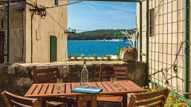 A wooden table with chairs, a bottle, and glasses, set against a stone wall with a view of the sea and a green gate.