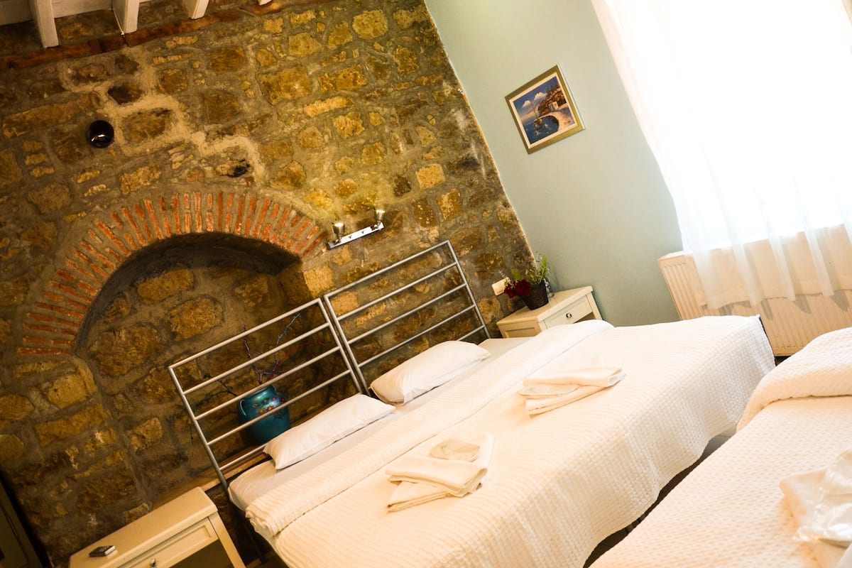 Una habitación de hotel con chimenea de piedra, dos camas con ropa blanca, una mesita de noche y un cuadro en la pared.