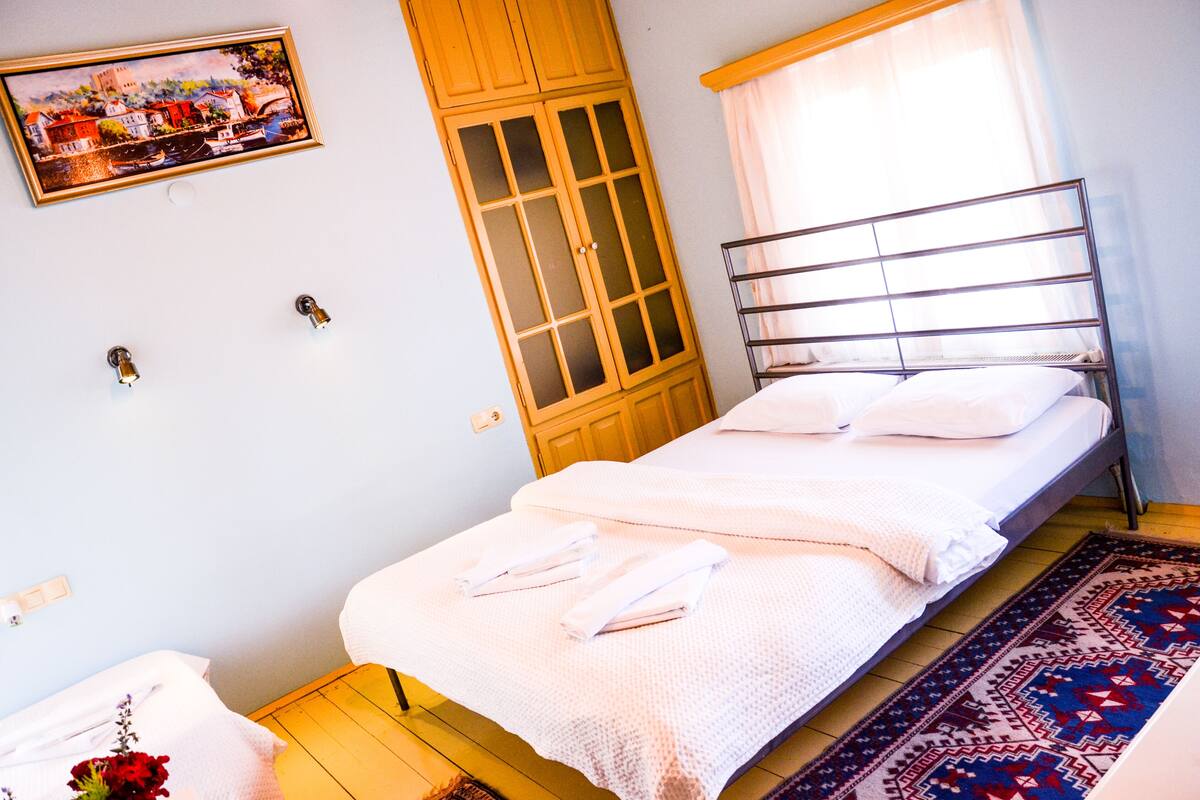 Una cama individual con ropa blanca, un armario de madera, una ventana con cortinas traslúcidas y una alfombra estampada en el suelo.