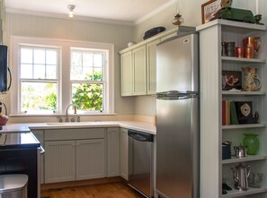 A kitchen with white cabinets, a stainless steel refrigerator, a dishwasher, and a window with a view of greenery.