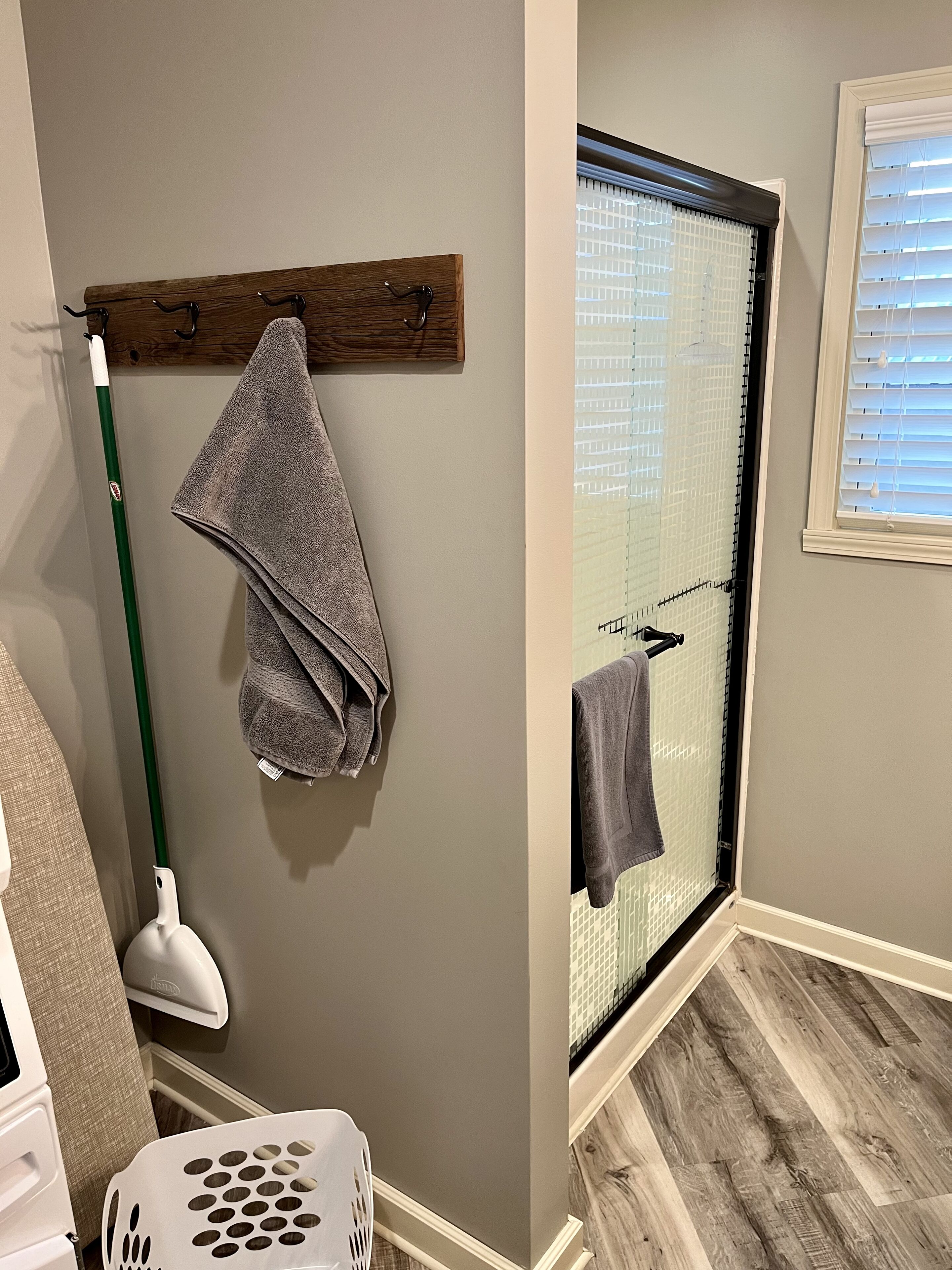 Un bagno con un portasciugamani in legno, uno straccio verde, un cesto per la biancheria bianco e una doccia con porta in vetro.