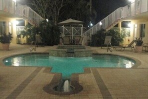 Una piscina con una fuente central, rodeada de sillas de descanso y plantas en macetas.
