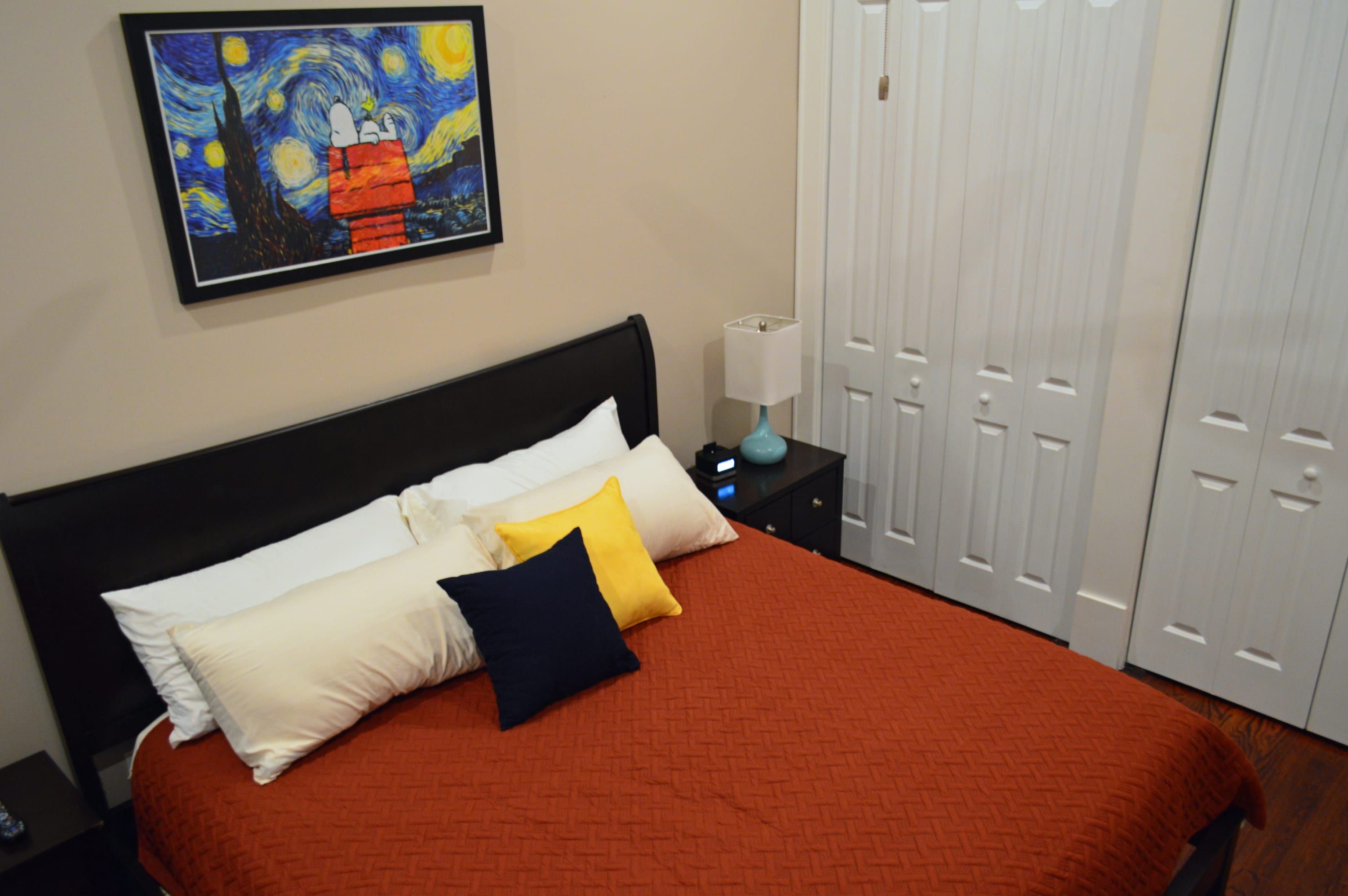 Un lit avec une couverture rouge, des oreillers blancs et bleu marine, et un tableau représentant un ciel nocturne étoilé.