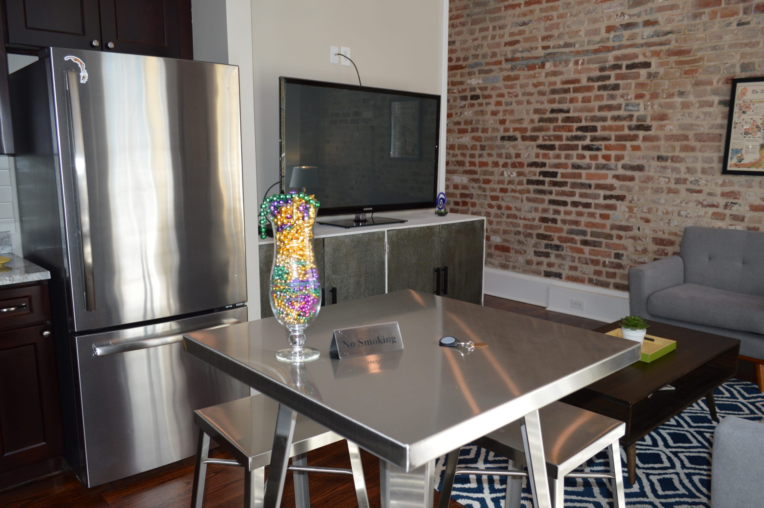 A modern kitchen with stainless steel appliances, a dining table with a 'No Smoking' sign, and a brick wall in the background.