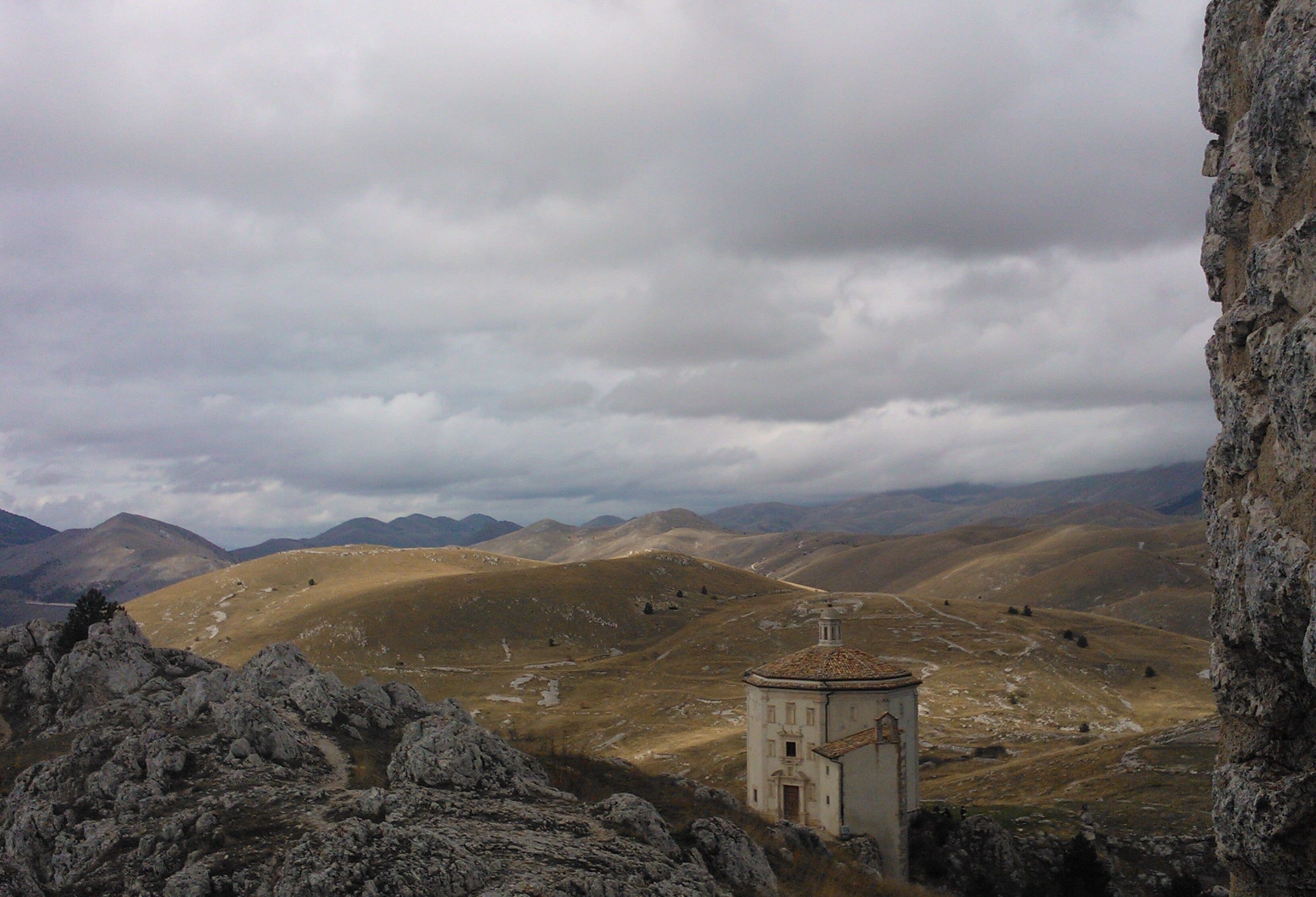 A mountainous landscape with a small building, possibly a chapel, surrounded by rocky terrain and distant hills.