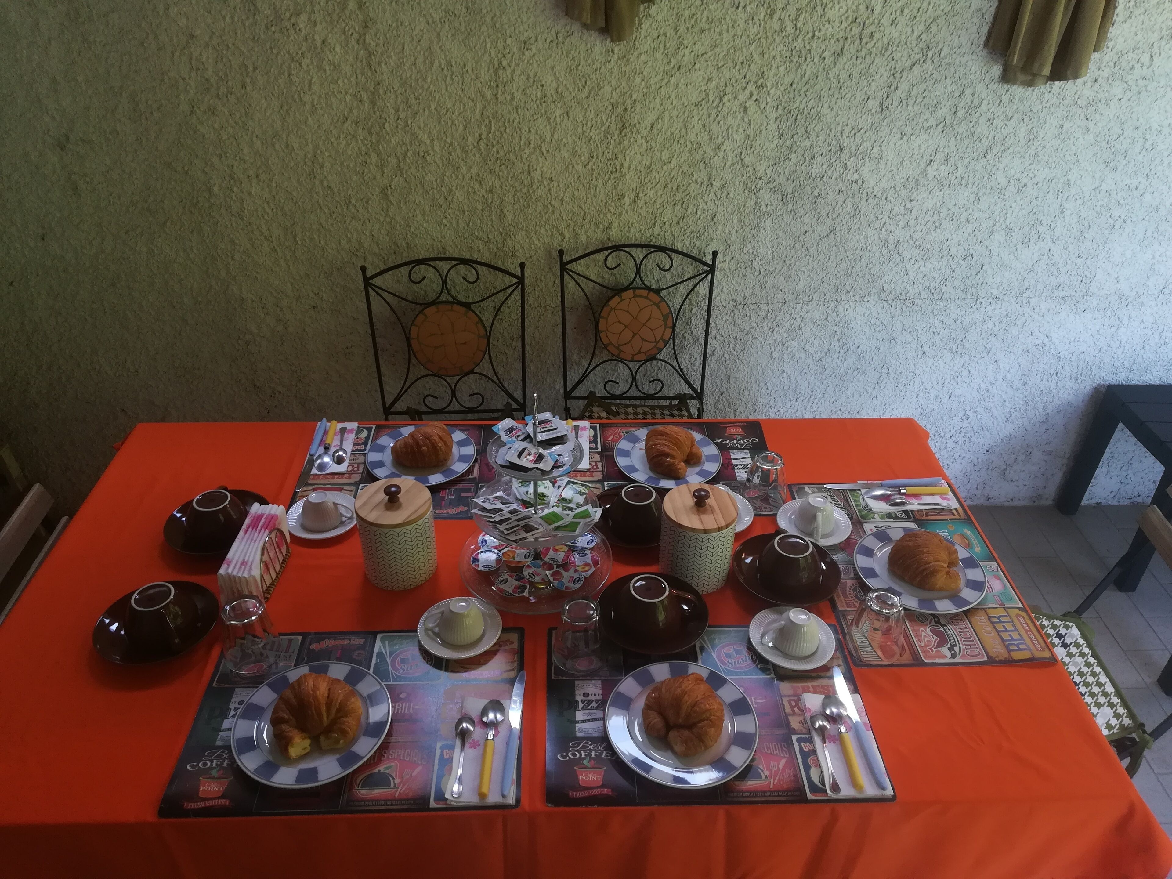 Une table dressée avec des croissants, du café et du thé, agrémentée d’un chemin de table à motifs et de chaises en métal.