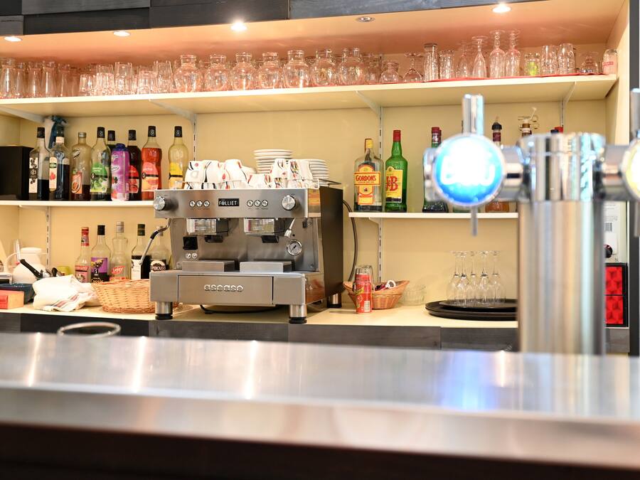 Zona de bar con una máquina de café, estantes con botellas y vasos.