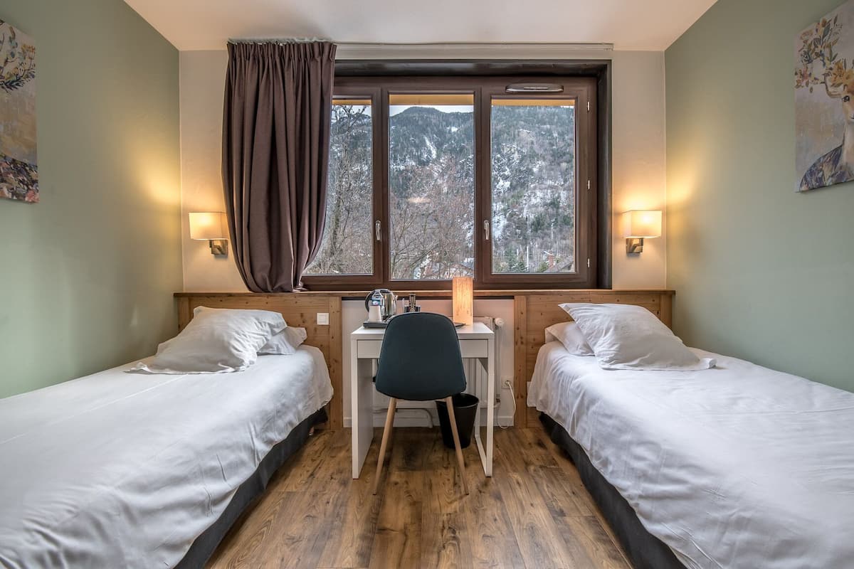 A room with two beds, a desk, and a chair. There is a window with a view of trees and mountains.