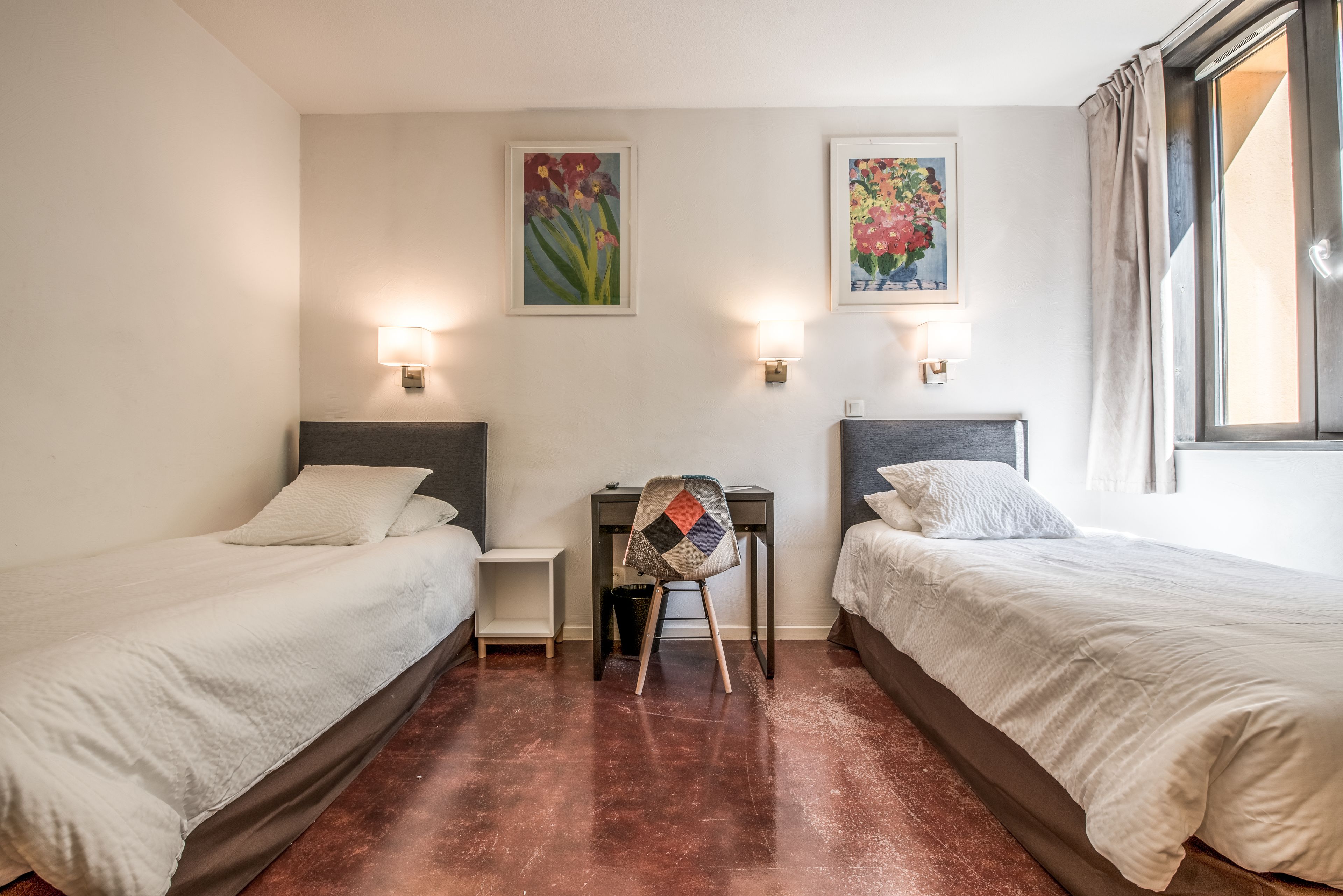Une chambre d’hôtel avec deux lits simples, un bureau et deux lampes fixées au mur.