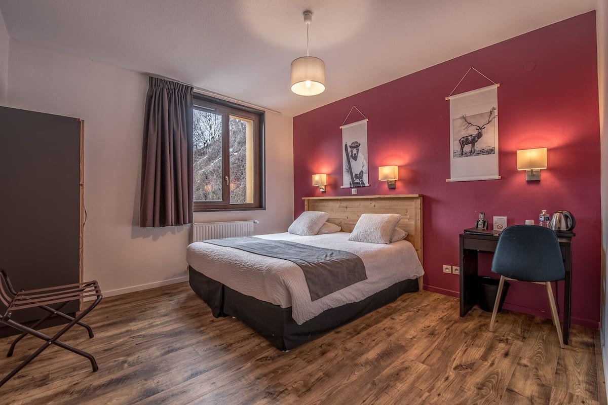A bedroom with a wooden floor, a bed with a grey and white duvet, a desk with a chair, and a window with curtains.