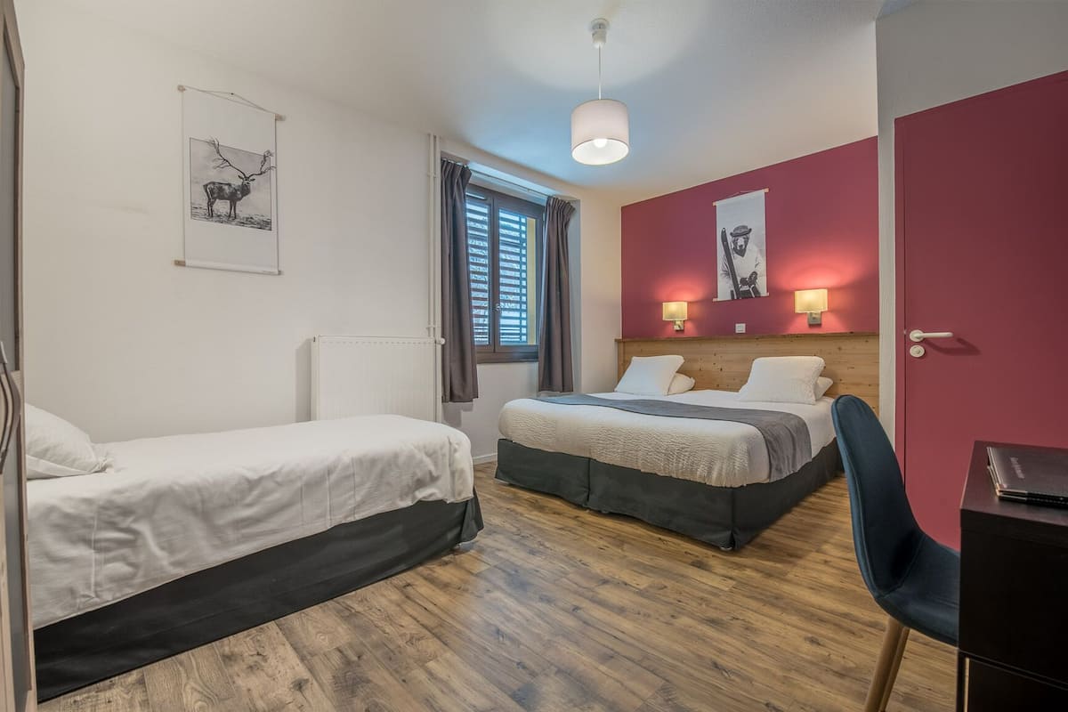 A room with two beds, a desk, and a chair. There are wall-mounted lamps and a window with blinds.