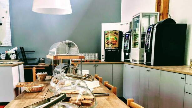 Un bufé de desayuno con gofres y otras pastas, una máquina de café, y una zona de estar con sillas de madera y una mesa.
