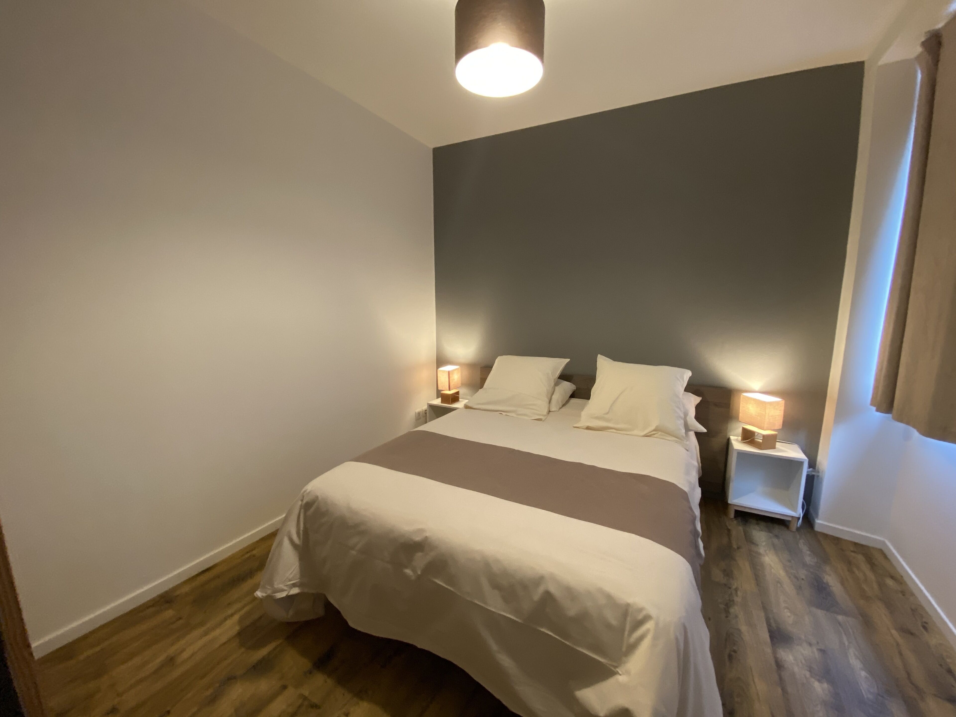 A neatly made bed with white linens and a brown accent, two bedside lamps, and a wooden floor.