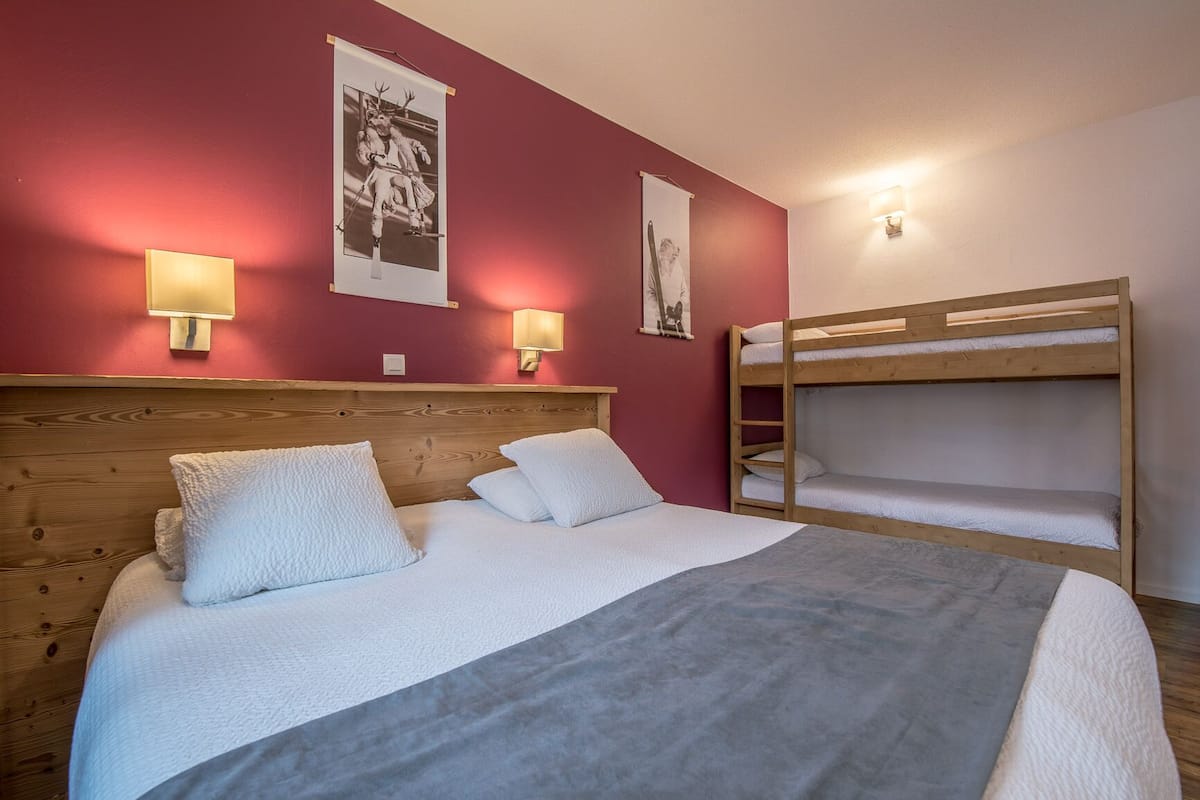 A room with a wooden bunk bed, a single bed with white bedding, and two wall-mounted lamps.