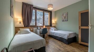 Quarto com duas camas de solteiro, uma escrivaninha, uma cadeira e janela com cortinas.