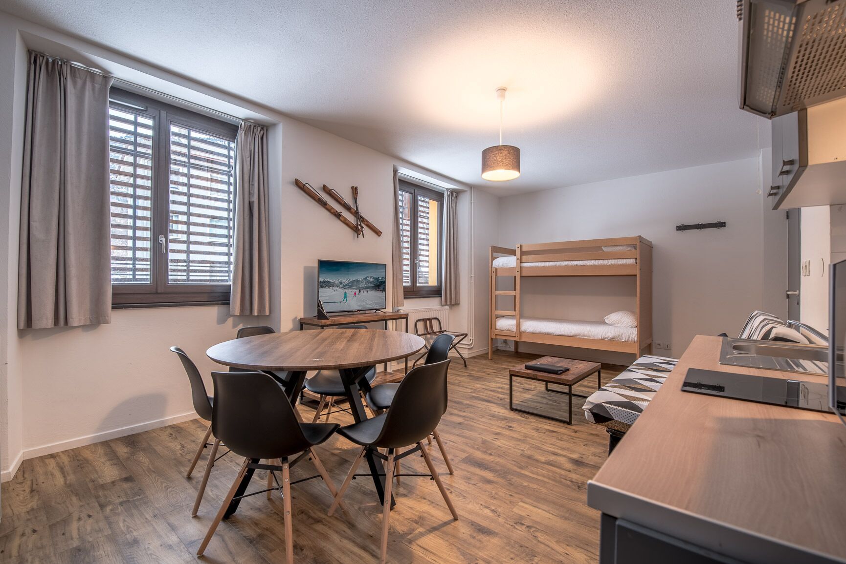 A compact room with a bunk bed, a dining table with chairs, a TV, and a large window with blinds.