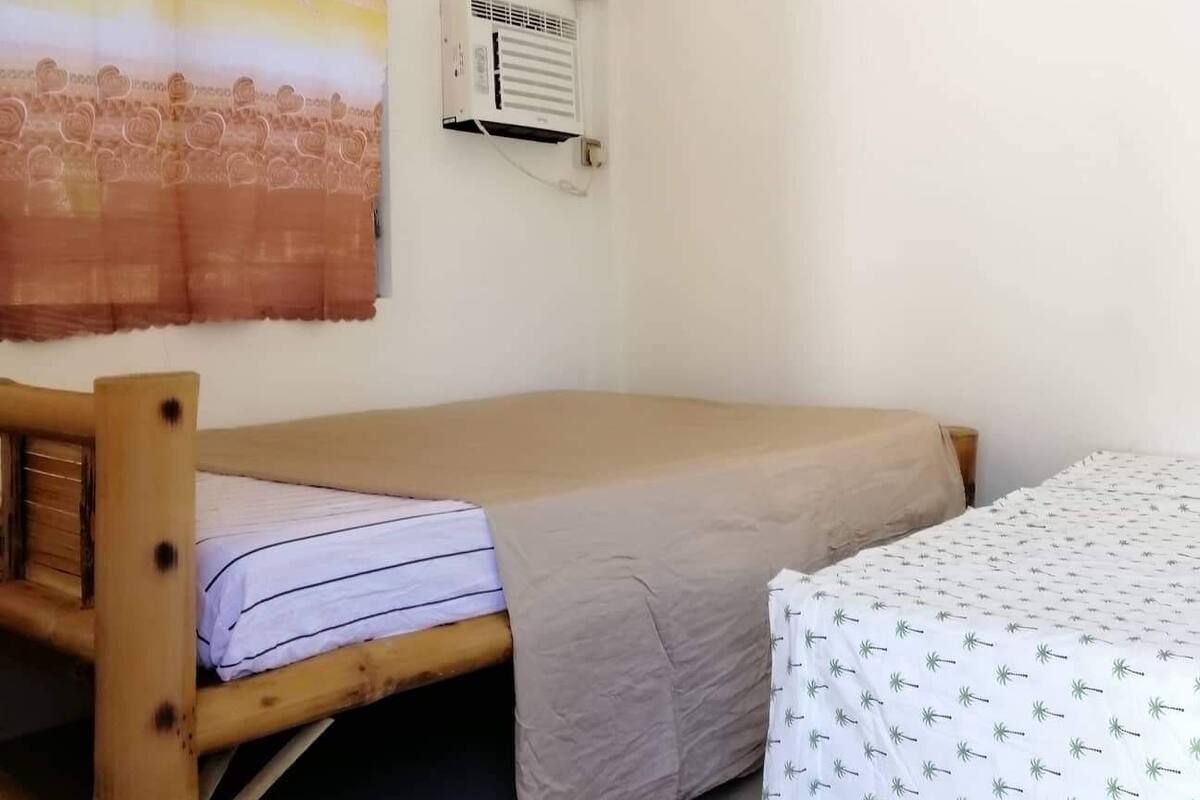 A small, single-room accommodation with a wooden bed, a small table, a wall-mounted air conditioner, and a clock.