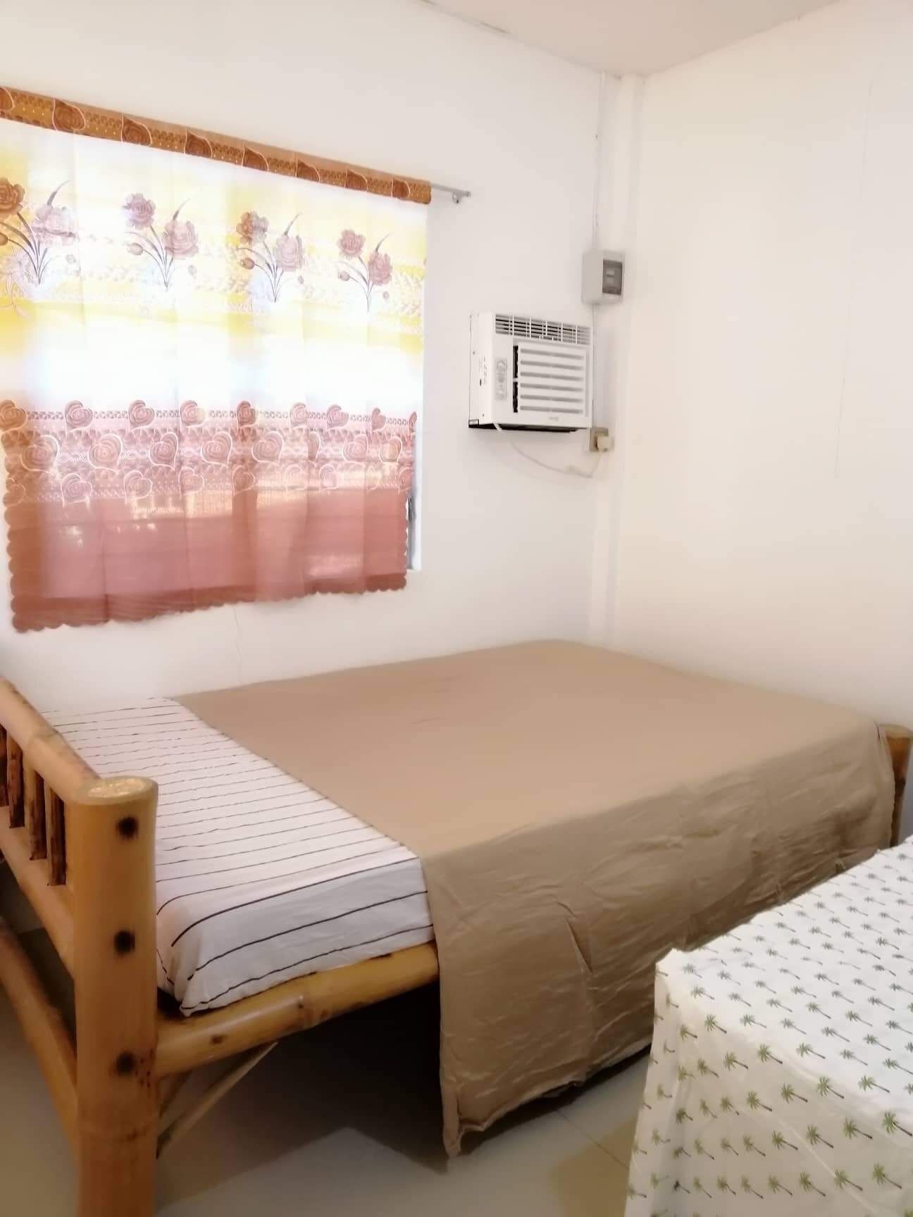 Una cama individual con estructura de madera, un colchón color beige y una sábana blanca estampada. Se observa una ventana con cortina floral y un equipo de aire acondicionado en la pared.