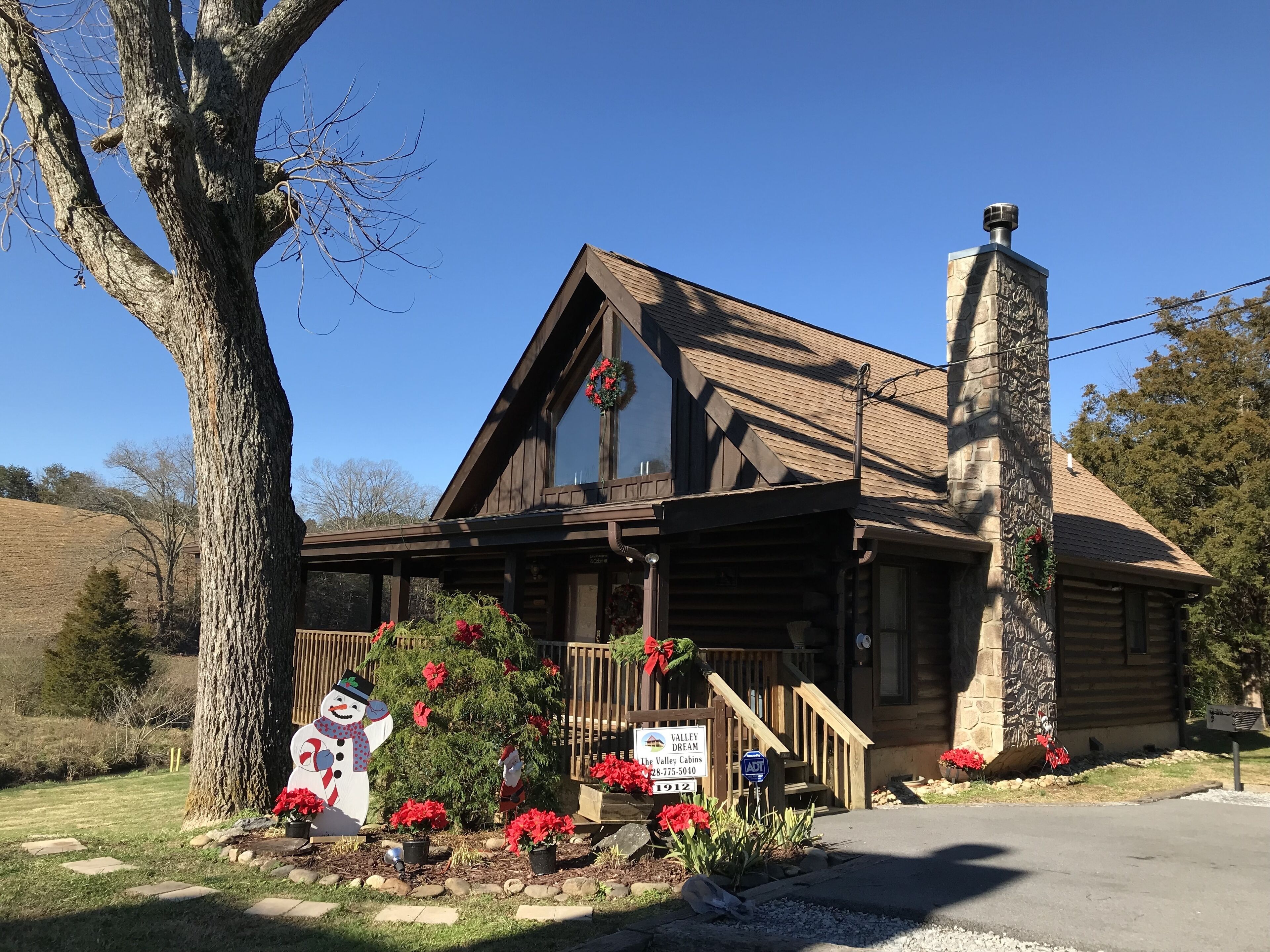 A log cabin with a stone chimney, a porch with red flowers, and a snowman decoration.
