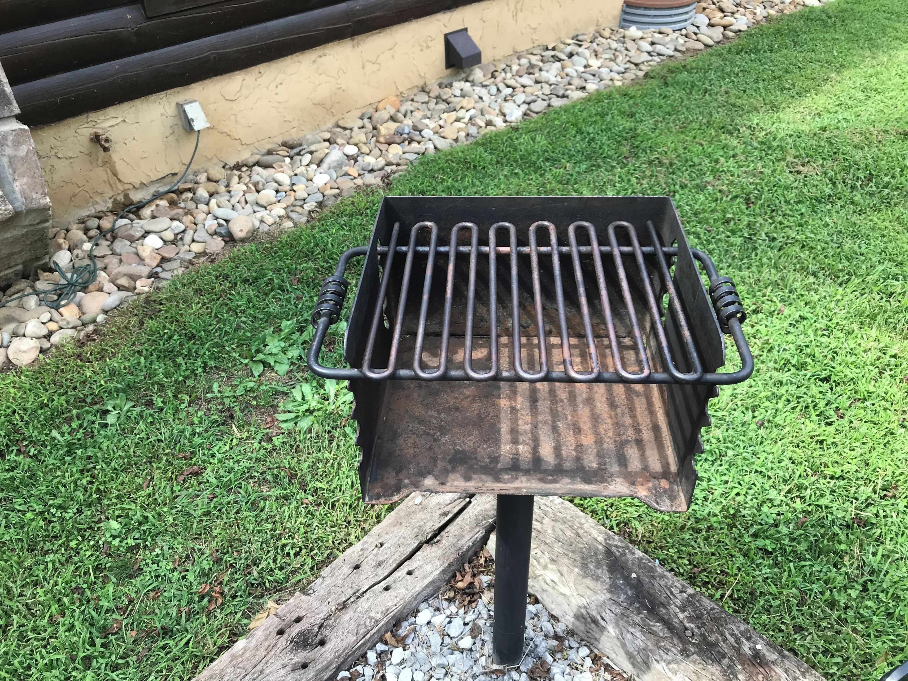 A metal grill with a wooden base, set on a grassy lawn with a stone border and a wooden fence in the background.