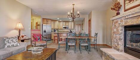 A living room with a fireplace, a dining area with chairs and a table, and a kitchen in the background.