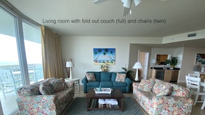 A living room with a fold-out couch, two matching chairs, a coffee table, and a view of the city through large windows. - Resort Style Amenities. NO extra admin fees added. Amazing parking spot!  (Orange Beach)