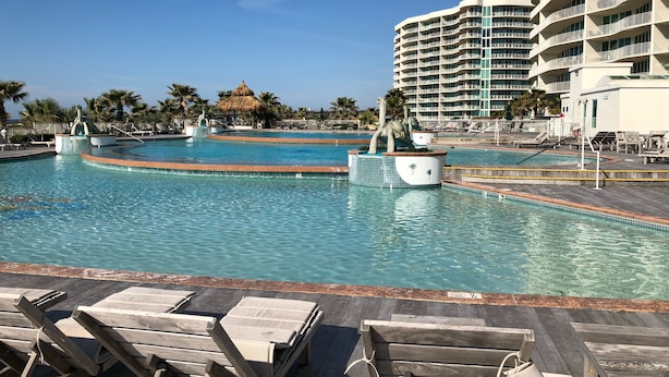 A resort pool with lounge chairs, a thatched-roof structure, and a multi-story building in the background. - Resort Style Amenities. NO extra admin fees added. Amazing parking spot!  (Orange Beach)
