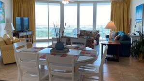 A spacious living room with a balcony, a television, and a dining area with a table and chairs. - Resort Style Amenities. NO extra admin fees added. Amazing parking spot!  (Orange Beach)
