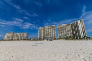 Un hotel frente a la playa con varios edificios altos, playa de arena y cielo azul despejado.