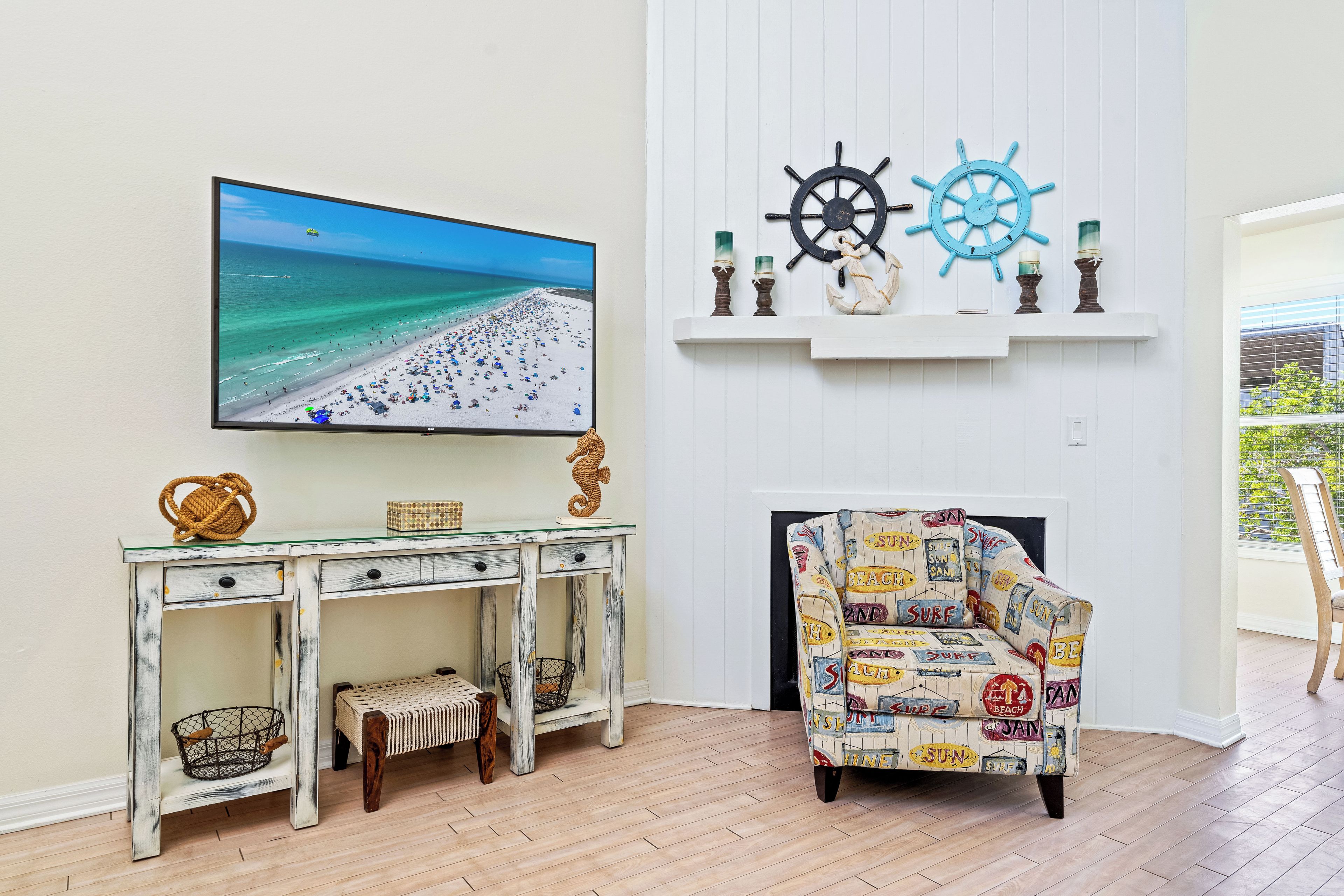 A living room with a beach scene TV, a white mantelpiece with nautical decor, a patterned armchair, and a rustic wooden table.