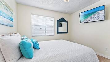 A bedroom with a bed, a ceiling fan, a window with blinds, a wall-mounted mirror, and a beach scene on the wall.