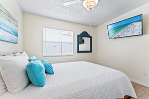 Un dormitorio con cama, ventilador de techo, ventana con persianas, espejo montado en la pared y una escena de playa en la pared.
