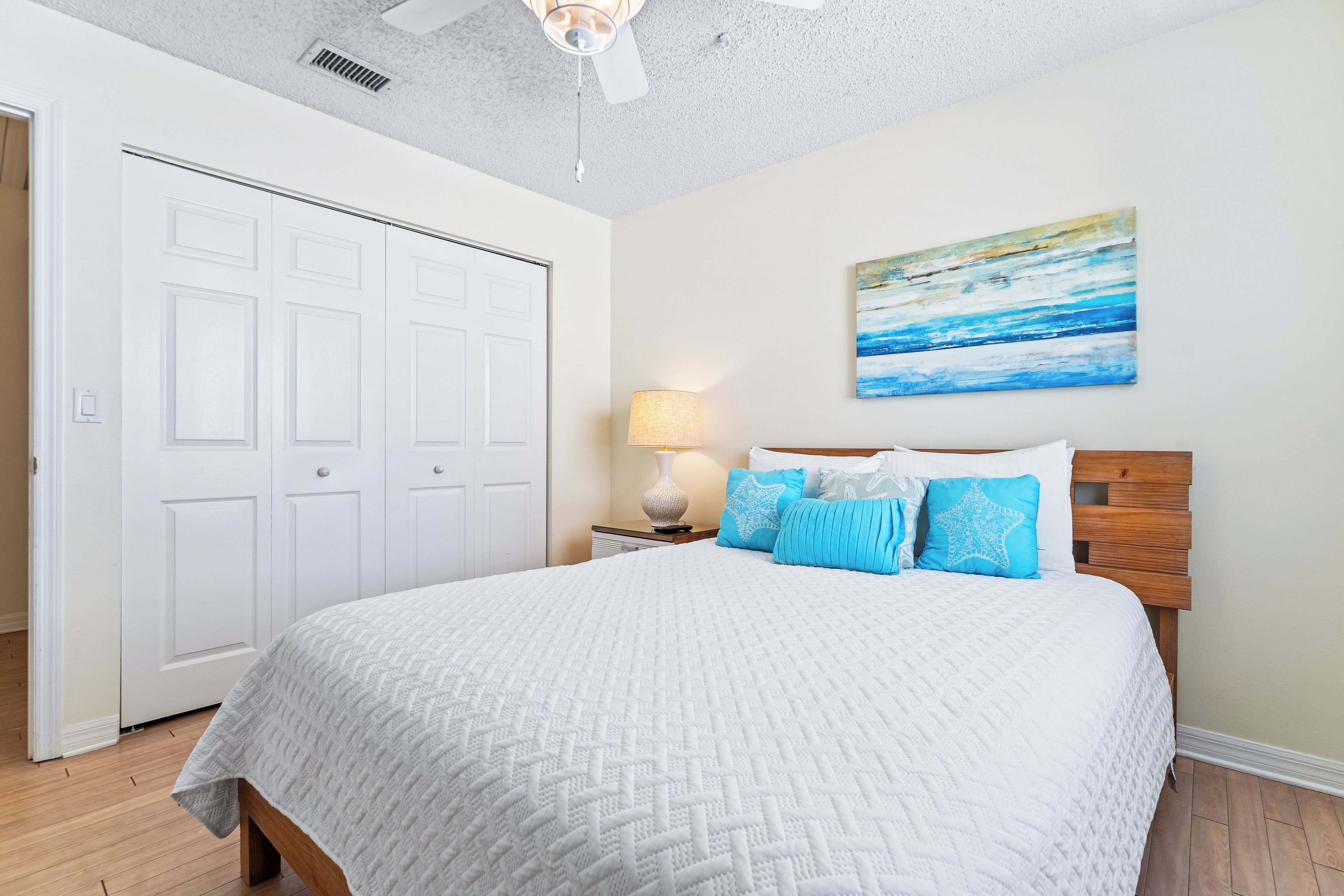 A bedroom with a large bed, a ceiling fan, a painting, and a nightstand with a lamp.